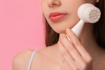Washing face. Young woman with cleansing brush on pink background, closeup