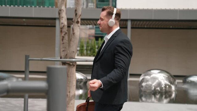 Happy project manager with headphone walking workplace while moving to relaxed music. Site view of smart caucasian businessman going to meeting while dancing and walking along the city street. Urbane.