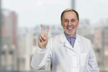 Cheerful senior caucasian doctor shows victory gesture sign. Blurred windows with cityscape view in the background.