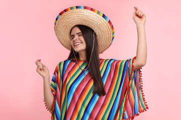 Young woman in Mexican sombrero hat and poncho dancing on pink background