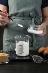Woman adding baking powder into measuring cup at black wooden table, closeup