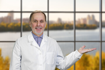 Happy smiling male senior doctor advertising a copy space. Checkered window background with landscape view.