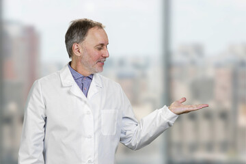 Happy male aged senior doctor advertising a copy space. Cheerful male psychisian standing indoors on blurred background.