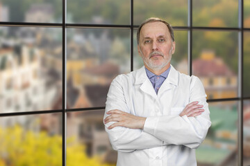 Portrait Of Happy Mature Male Doctor With Folded Arms. Checkered windows backgorund with cityscape view.