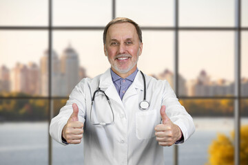 Happy smiling senior doctor shows both thumbs up. Checkered window background with cityscape view.