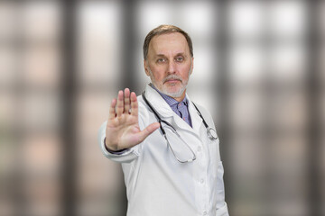 Serious physician doctor shows stop gesture sign. Elder male doctor shows his palm. Blurred checkered windows background.