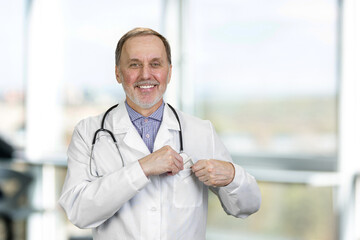 Portrait of happy senior male doctor puts money in his pocket. Smiling aged physician indoors.