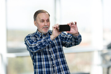 Handsome smiling senior man taking a photo on his smartphone. Standing in a bright office blurred windows background.