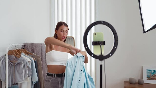 Young social medial content creator woman make fashion video. Blogger smiles to camera using selfie stick and light ring while making persuasive online clothing sell vlog to audience or follower. Adit