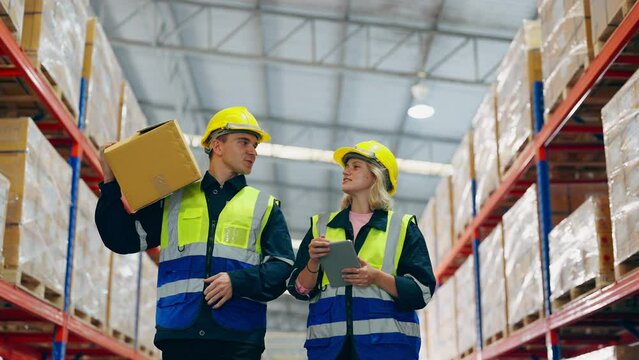 Young man and woman warehouse worker wearing helmet and vest checking order details on tablet computer for goods and supplies on shelves in storehouse