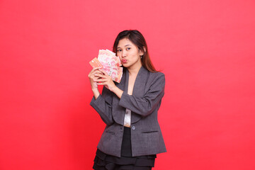 The expression of a cheerful Asian girl director with both hands holding a credit card, debit card and money kissing the camera wearing a gray jacket and skirt on a red background. for advertising