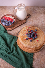 A plate of golden pancakes topped with blueberries, accompanied by a bowl of sour cream, a portion of fresh cottage cheese and a glass of milk. On a beige-golden background with a green runner.