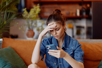 Stressed woman sitting on couch looking at phone with head in hands in living room