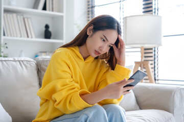 Bad news on device screen. Upset asian woman frustrated by problem with work or relationships, sitting on couch, feeling despair and anxiety, loneliness, having psychological trouble