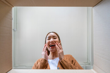 Low angle view of surprised young asian woman unpacking. Opening carton box and looking inside. Packaging box, delivery service. Human emotions and facial expression