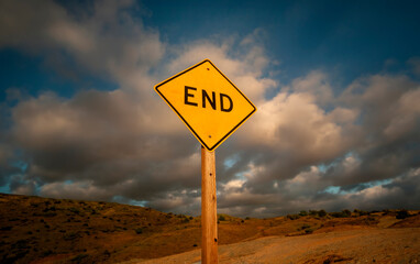 Yellow diamond shaped traffic sign on a dirt road with the word End written on it against a stormy cloudy sky
