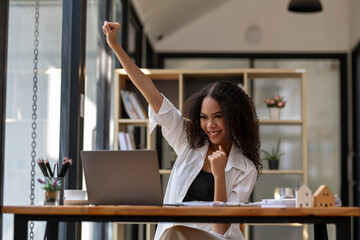 Ecstatic businesswoman with curly hair cheering at her desk after receiving positive news on her...