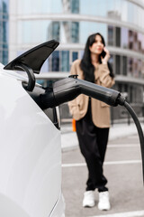 A business woman uses an electric charging station for her modern car against the backdrop of a modern city. Concept of economic charging and modern technologies.
