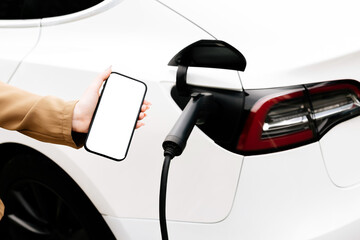 Close-up photo of an electric car charging. A woman holds a phone next to her with a white screen for your text. Concept of modern electric vehicles, technology and energy.