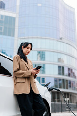 Obraz premium A businesswoman is waiting to charge her electric car in the parking lot of a business center with a phone in her hands. Futuristic eco-friendly electric car uses alternative energy in urban lifestyle