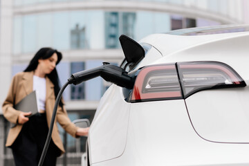 A business woman uses an electric charging station for her modern car against the backdrop of a...