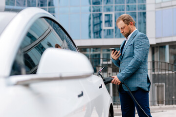 A middle-aged businessman charges an electric car from a charging station powered by renewable...
