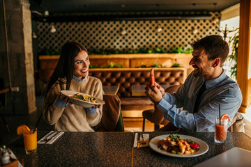 Happy young woman holding a dish during the lunch and photographing with her boyfriend in a...