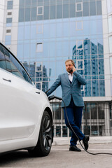 A young successful businessman in a stylish suit uses a telephone against the backdrop of a modern business center and a modern electric machine. Concept of electric vehicles, business and style.