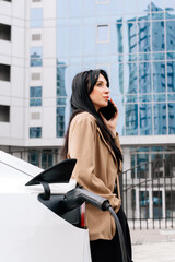 Close-up of a modern electric car charging against the backdrop of a business woman and a modern business center. Concept of using energy, electrical machines and modern technologies.