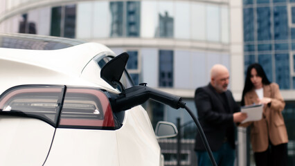 An unrecognizable modern electric car is charging. In the background, two business people, a man...
