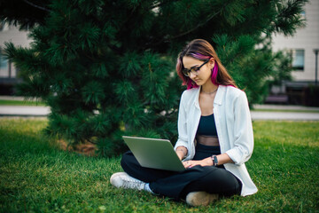 Pretty long-haired college girl with laptop preparing for exam while sitting on campus lawn. Charming young woman student in white shirt studying outside class in the park.