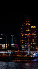 Fototapeta premium Millennium Bridge illuminated at night with white lights reflecting its lights on the River Thames and its bank, with skyscrapers illuminated behind and part of the London Eye illuminated with purple