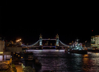 Obraz premium Front view of Tower Bridge illuminated at night and the war museum ship HMS Belfast docked on the River Thames with the lights of restaurants, buildings reflected in the water.