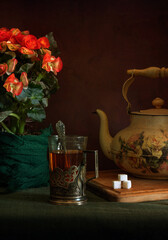 Hot, aromatic tea in a glass with a glass holder, an old teapot and a blooming begonia.