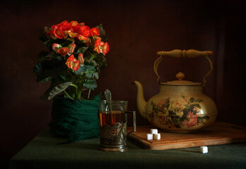 Hot, aromatic tea in a glass with a glass holder, an old teapot and a blooming begonia.