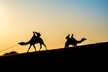 Silhouette of camel with two people sitting on it crossing over sand dunes in Sam Jaisalmer...
