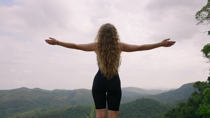 Curly-haired woman embraces mountain breeze, arms wide open. Feels freedom, peaceful nature surround. Hikes, enjoys scenic view, wellness retreat. Moment of joy, empowerment in green hills. Slowmo