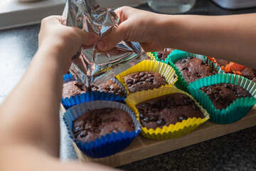 Child hands spreading chocolate icing onto chocolate cupcakes with chocolate chips sitting inside colorful baking liners