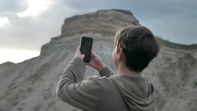 A cute Caucasian teenager takes pictures of himself using a smartphone camera with his arm outstretched. The boy will take a selfie in the mountains at sunset. Active lifestyle