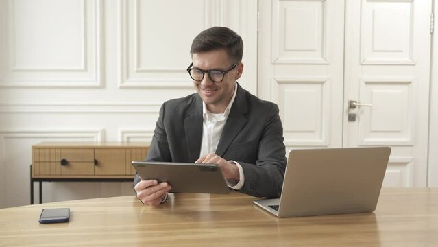 A business professional works in an office, wearing a suit and using a laptop and tablet computer for their workstation.
