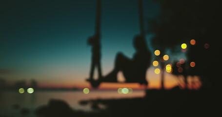 A person sits on a swing in a blurred capture, sunset in Thailand. The motion blur adds a sense of...
