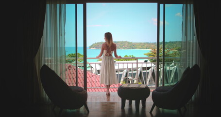 Young woman in white dress stand on balcony enjoy ocean view. Female relax in Phuket, Thailand...
