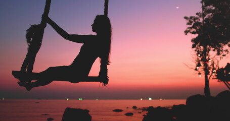 Woman silhouette sway wooden rope swing on beach, bright pink blue sunset. Vacation beautiful...