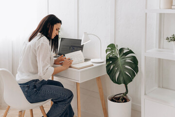 Young brunette freelancer woman sitting at desk using laptop writing notes while studying online, watching webinar, looking at pc screen learning web classes or remote working from home. Side view