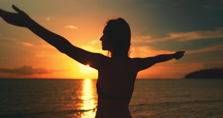 A woman standing on Koh Ma Beach in Koh Phangan Island, Thailand, during sunset, with the sun setting over the horizon and casting a warm glow on the sandy beach. Raise hands up enjoy holiday vacation