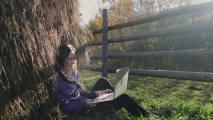 A woman with long hair sits peacefully in the lush green grass, typing on laptop under a stack of hay. Freelancer work remotely in autumn countryside at sunset. Travel, freedom, active lifestyle