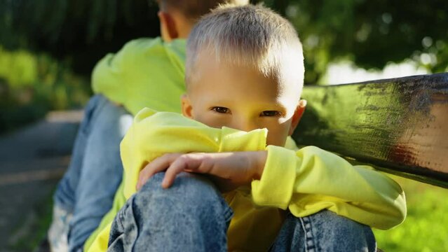 Brothers sit back-to-back on park bench after dispute. Young boy in yellow hoody rests his chin on his hands, looking thoughtfully at camera with hint of sadness, following argument with his sibling