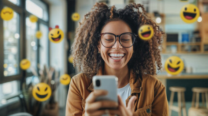 A young smiling black woman holding a mobile phone in the office with emoji smiles flying around her. The person receives a pleasant message - Powered by Adobe