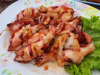 A white plate with a grilled squid resting on a bed of green lettuce. The squid is long and white, with grill marks and visible tentacles.