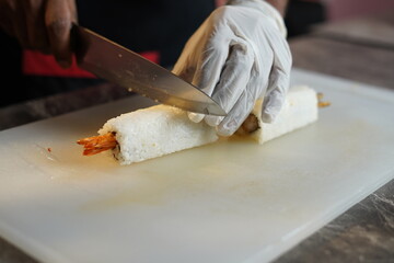 Japanese sushi is cutted and plated by gloves wearing hand chef with sharp knife, shrimp and sauces are visible in between sticky rice and bamboo role and firing the sushi role with burner 